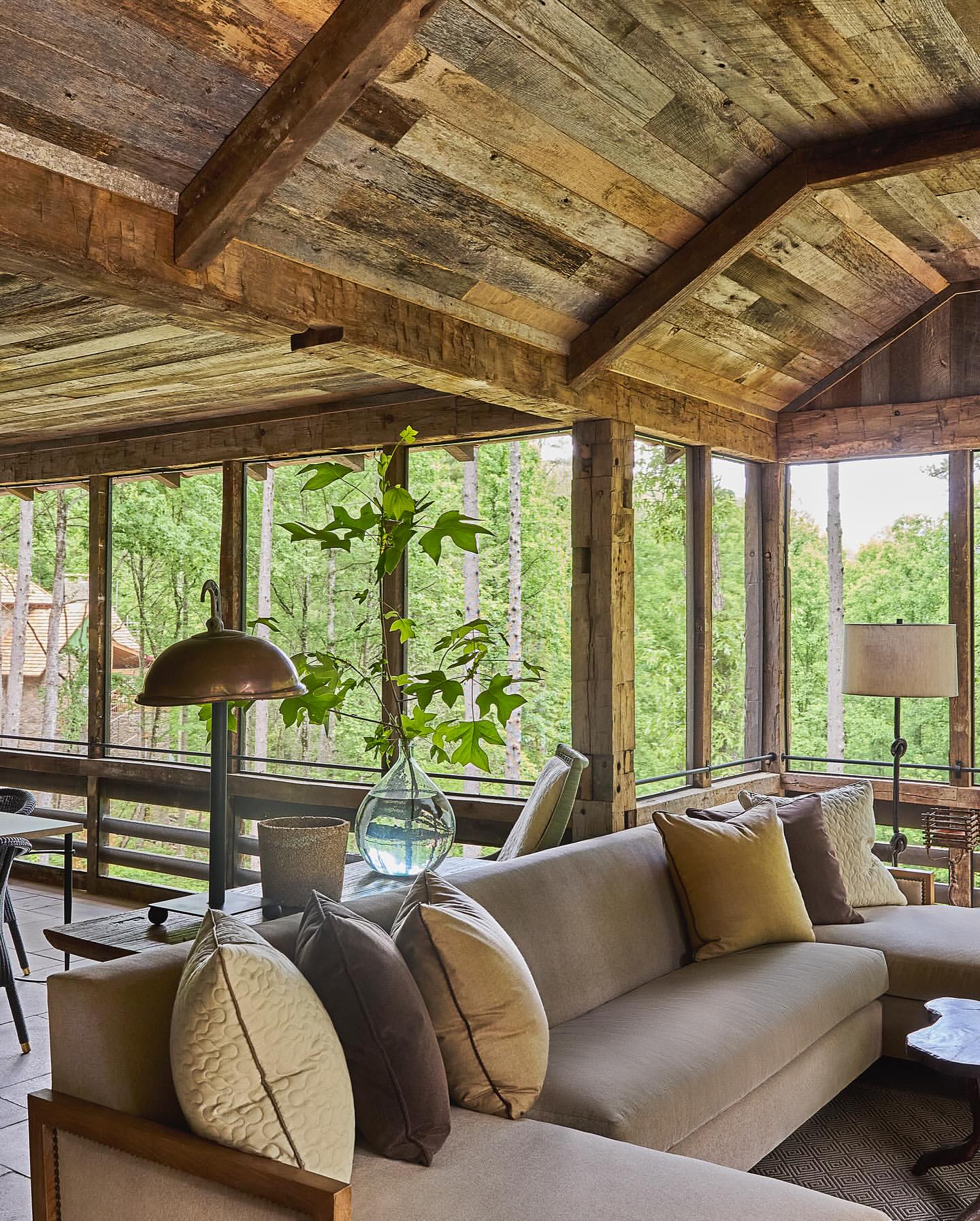 Cozy indoor sunroom featuring a neutral sectional sofa with decorative pillows, a glass vase with greenery, and rustic wood paneling.