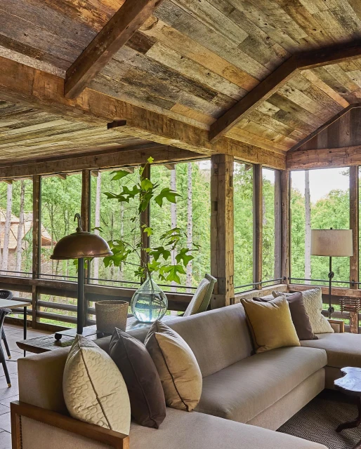Cozy indoor sunroom featuring a neutral sectional sofa with decorative pillows, a glass vase with greenery, and rustic wood paneling.