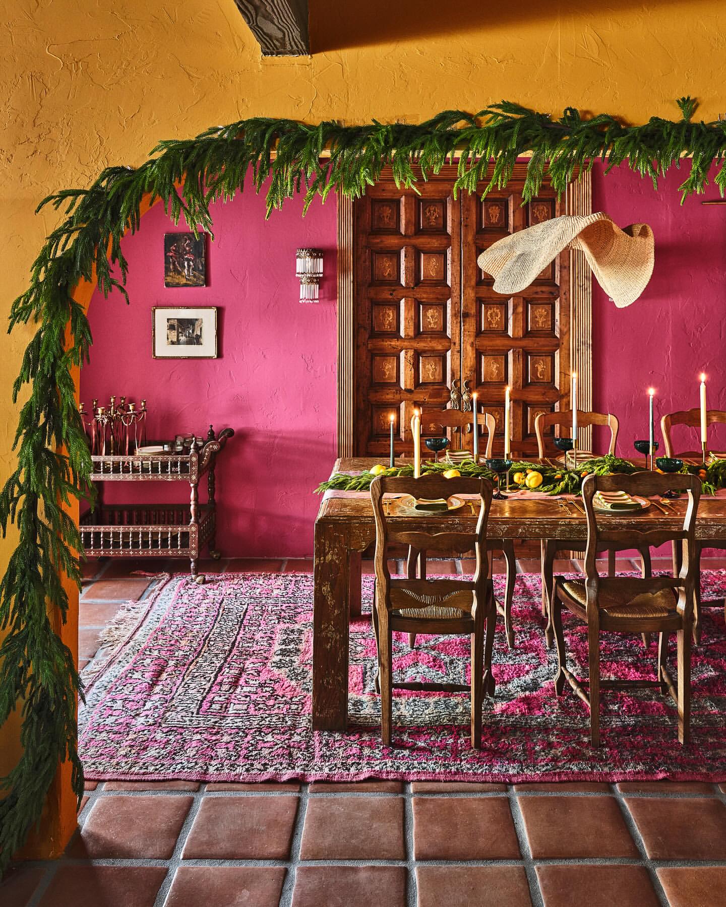 Warm-toned interior featuring a rustic wood table set for a meal, surrounded by wooden chairs, pink walls, and a terracotta tile floor.