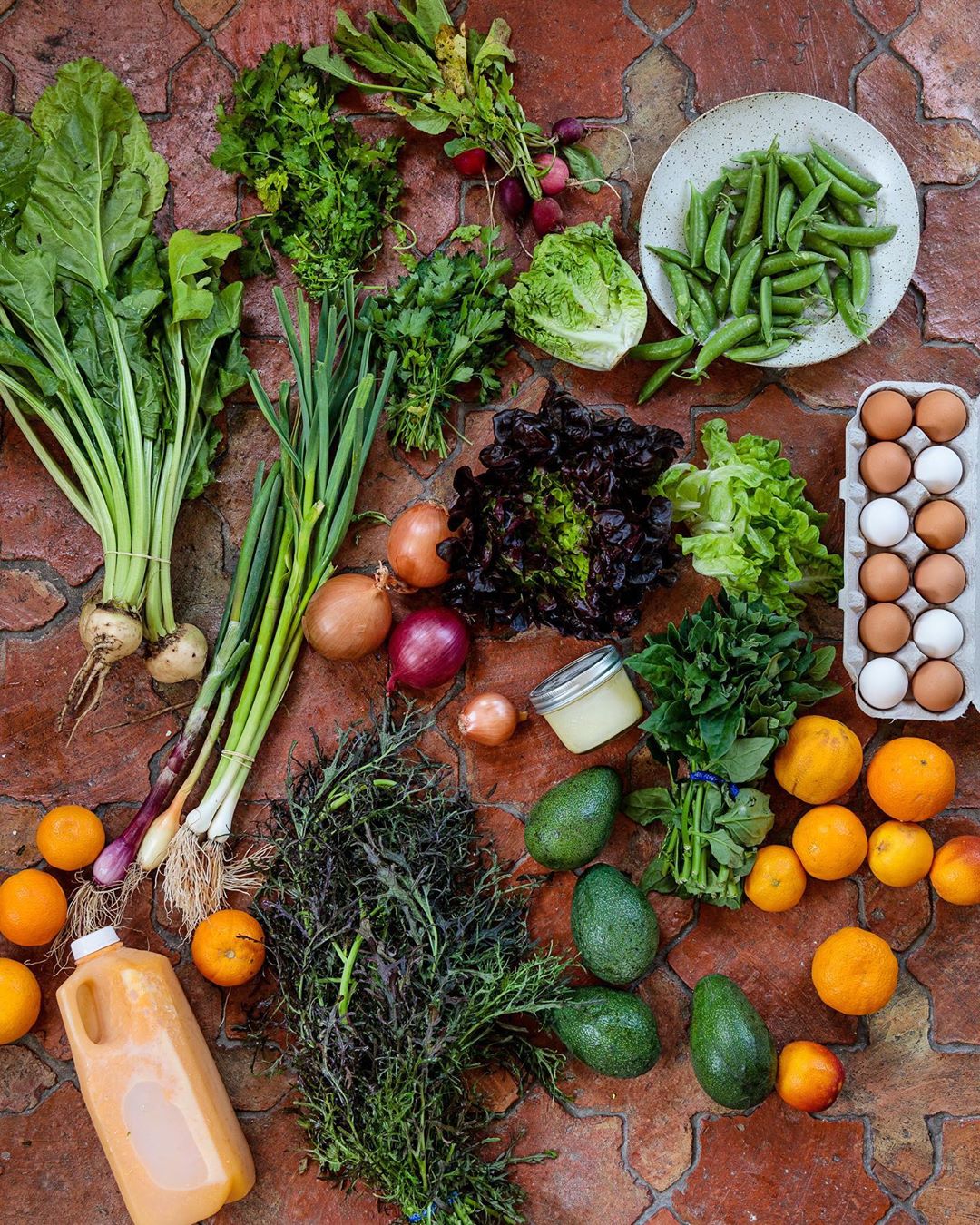 Overhead shot of fresh produce: lettuce, greens, onions, avocados, oranges, eggs, juice on a brick surface in natural light.