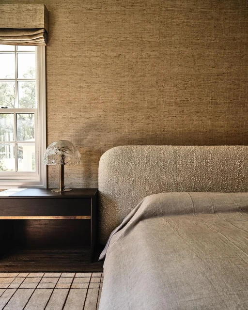 Bedroom interior with a neutral-toned upholstered bed, dark wood nightstand with a lamp, window with shade, and patterned wallpaper.