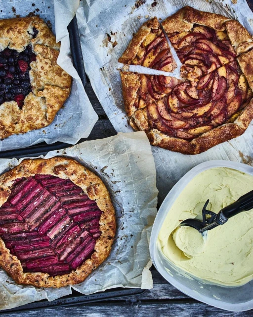 Overhead shot of three rustic galettes—blueberry, peach, and rhubarb—alongside a container of ice cream on a weathered wooden surface.