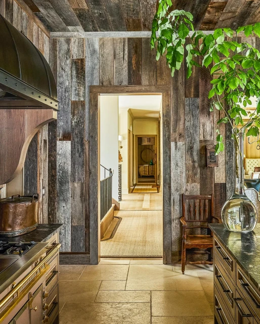 Rustic kitchen interior with wooden walls and ceiling, stone floors, island with dark countertop, and hanging plant in a home.