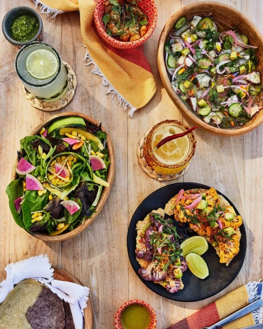 Overhead shot featuring a wooden table arrangement of food dishes, including salads, tostadas, and drinks with limes, creating a culinary scene.