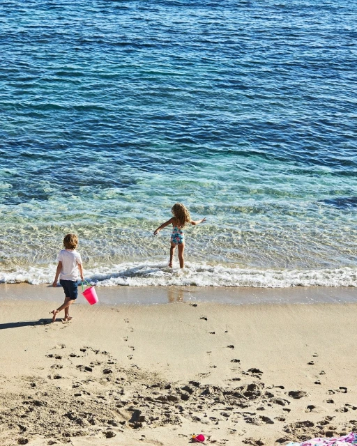 Two young children play in the shallow surf on a sunny beach; one child carries a pink bucket, with blue ocean in background.