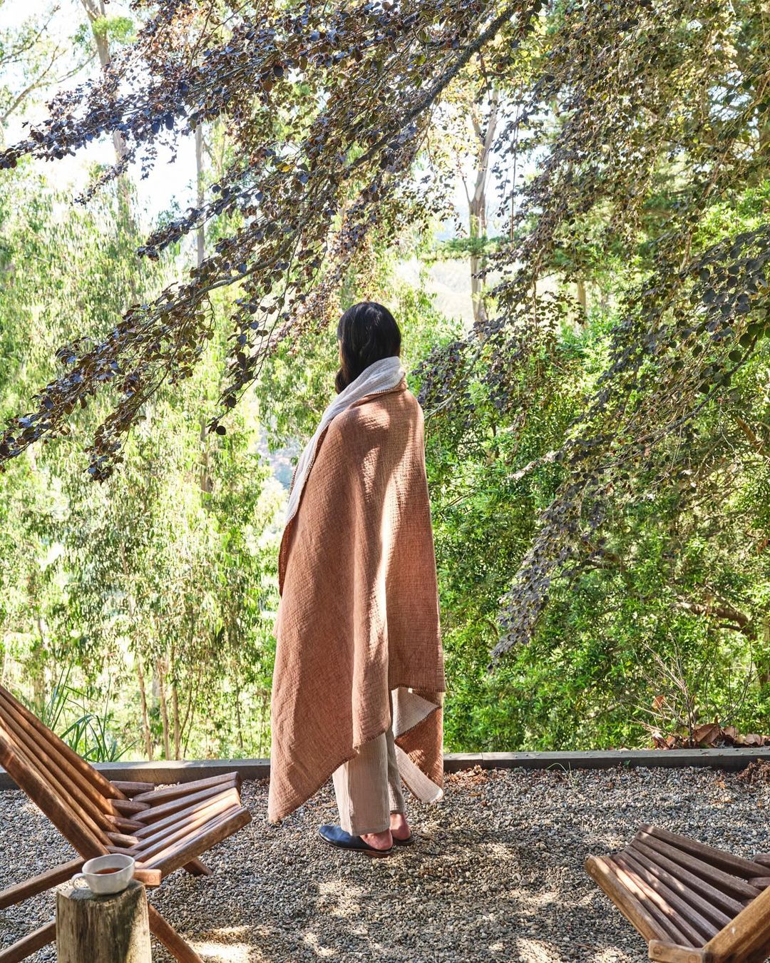 Woman wrapped in a tan blanket, standing on a patio overlooking a forest. Wooden lounge chairs and a cup add to the serene scene.