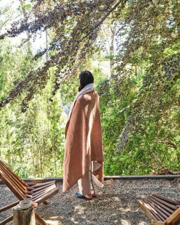 Woman wrapped in a tan blanket, standing on a patio overlooking a forest. Wooden lounge chairs and a cup add to the serene scene.