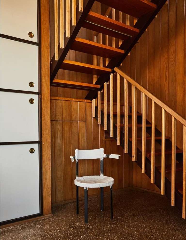 Interior view featuring a white chair against a wooden wall with staircase. Room is lit with natural light highlighting wood textures.