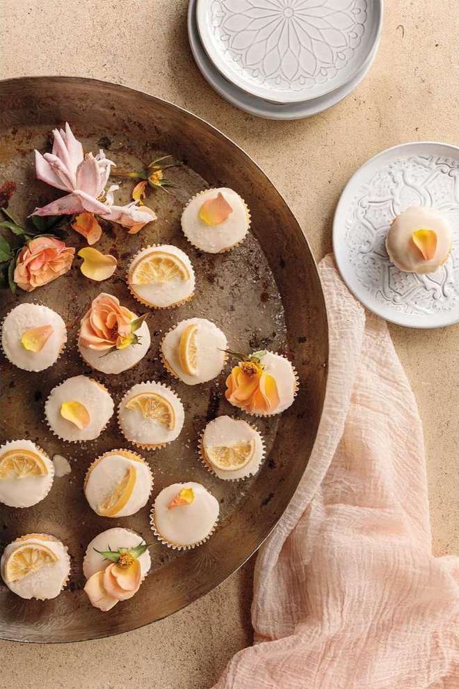 Overhead shot of mini-cupcakes decorated with orange slices and flower petals, arranged on an old metal tray with peach-colored cloth.