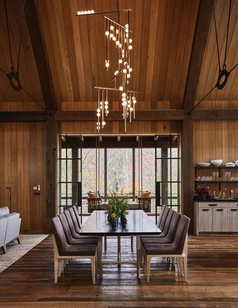 A modern dining room features a long dining table surrounded by upholstered chairs, beneath an elaborate chandelier and wood paneled walls.