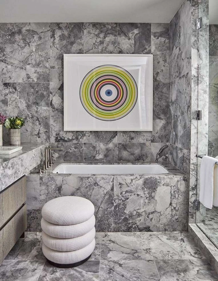 Luxurious bathroom interior featuring grey marble tile walls, a built-in bathtub, a white round ottoman, and a colourful circular abstract artwork