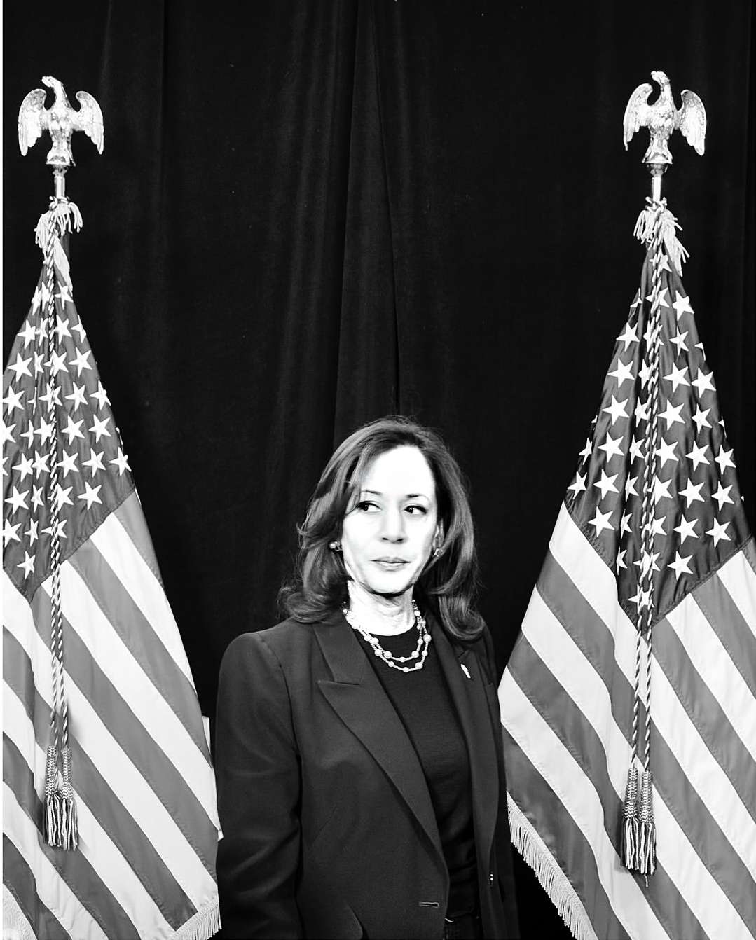 Black and white portrait of a woman in a dark suit jacket standing in front of two American flags with black background.