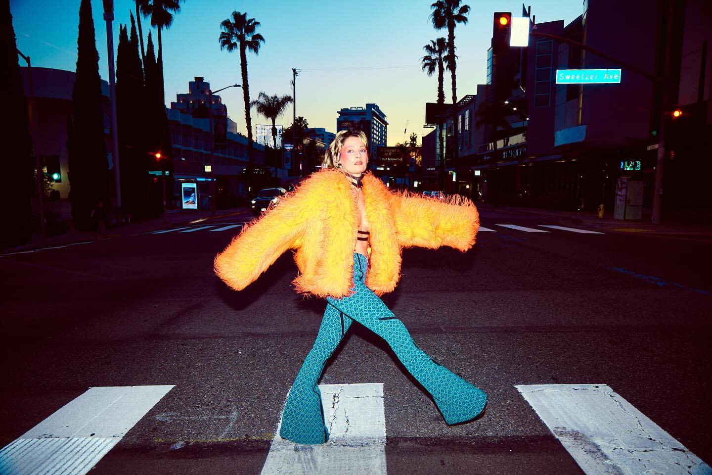 Woman in bright orange jacket strides across crosswalk at dusk in a city with palm trees and building silhouettes in the background.
