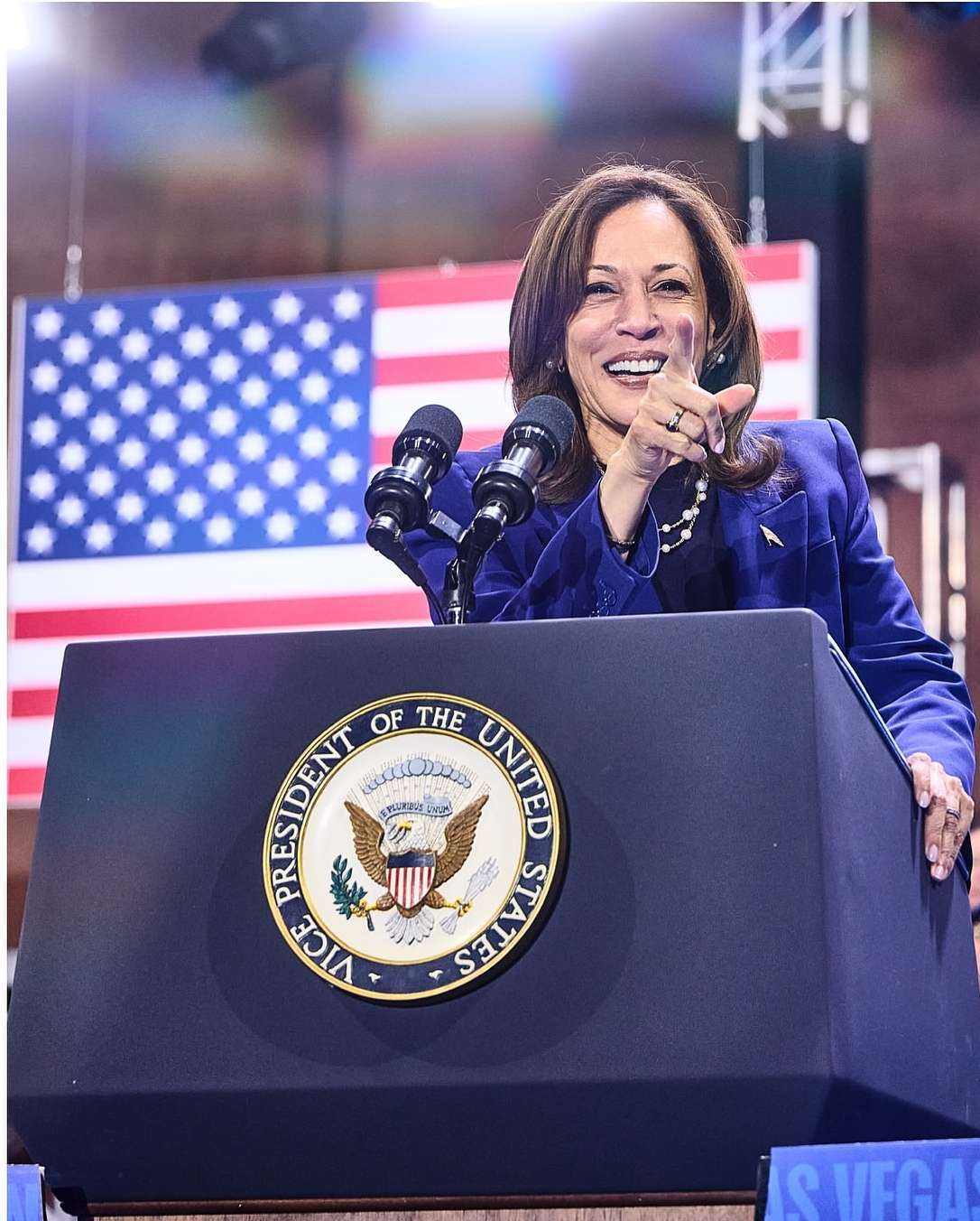 Kamala Harris stands at a podium, smiling and gesturing at a crowd. An American flag is visible in the background behind her.