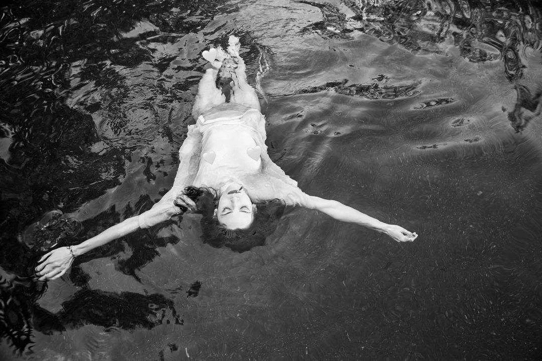 Woman in light dress floats serenely in dark water, arms outstretched, captured in a black and white photograph from above.