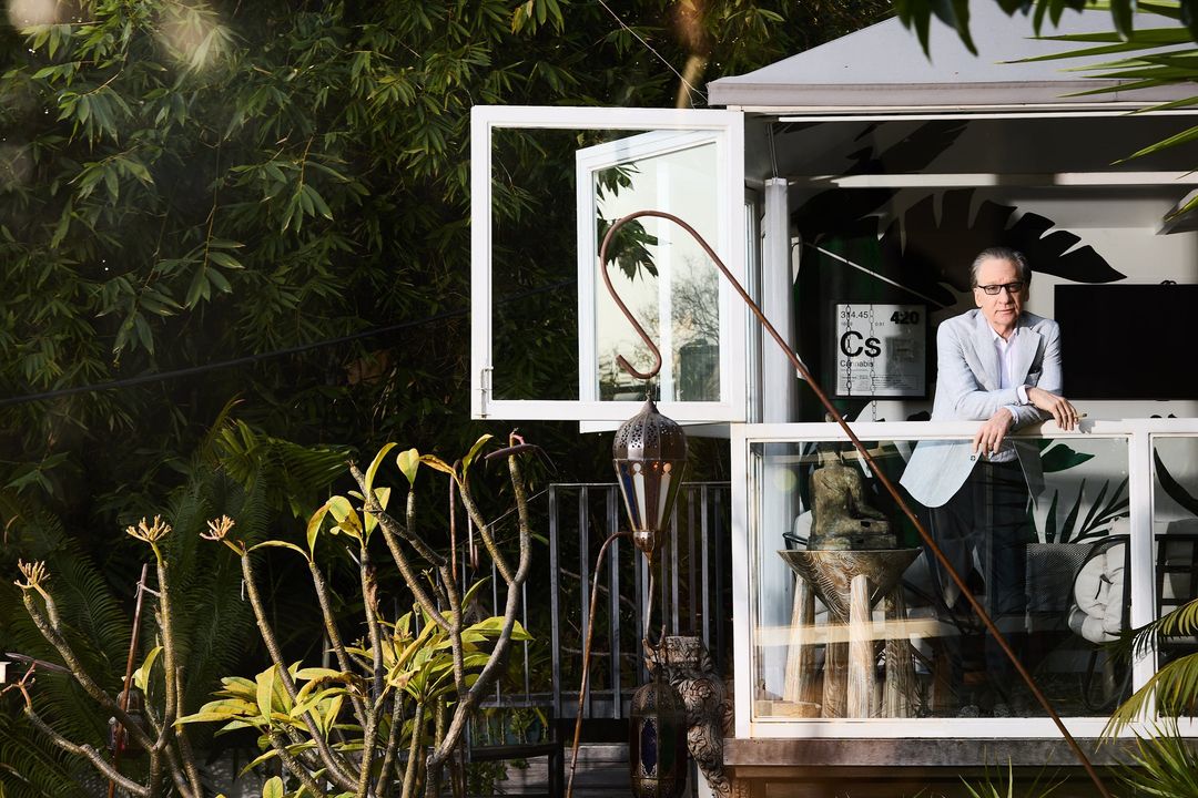 Adult man stands on balcony of modern home surrounded by tropical greenery, wearing blue blazer and glasses, overlooking garden.
