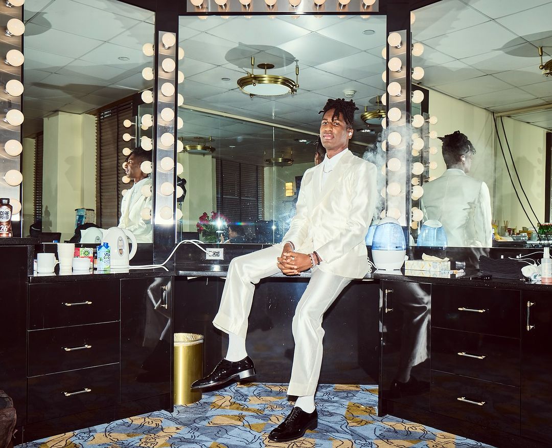 Stylish man in a white suit sits on a black vanity with bulbs surrounding the mirror in an indoor setting with patterned carpet.