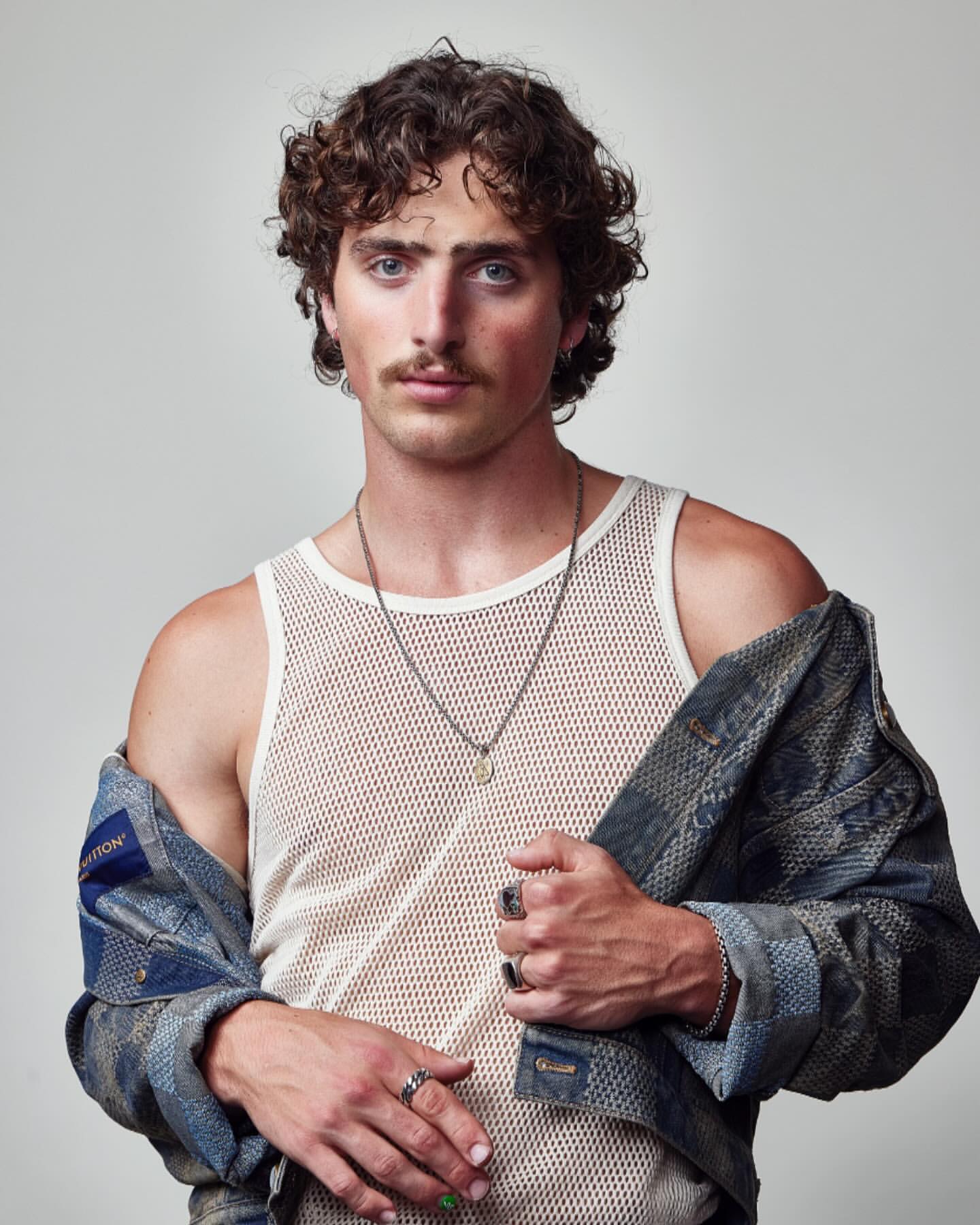 Indoor portrait of a young adult male with curly brown hair, wearing a mesh tank top and denim jacket against a neutral backdrop.