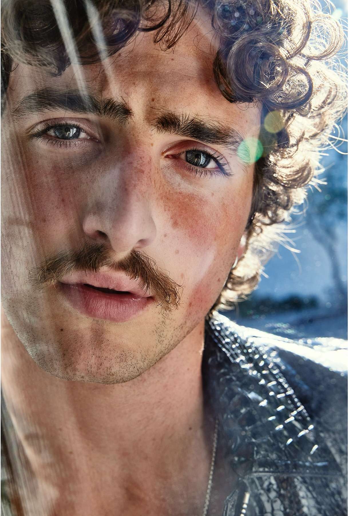 Close-up portrait of a young adult male with curly hair, a mustache, and a textured black jacket looking directly at the camera indoors.