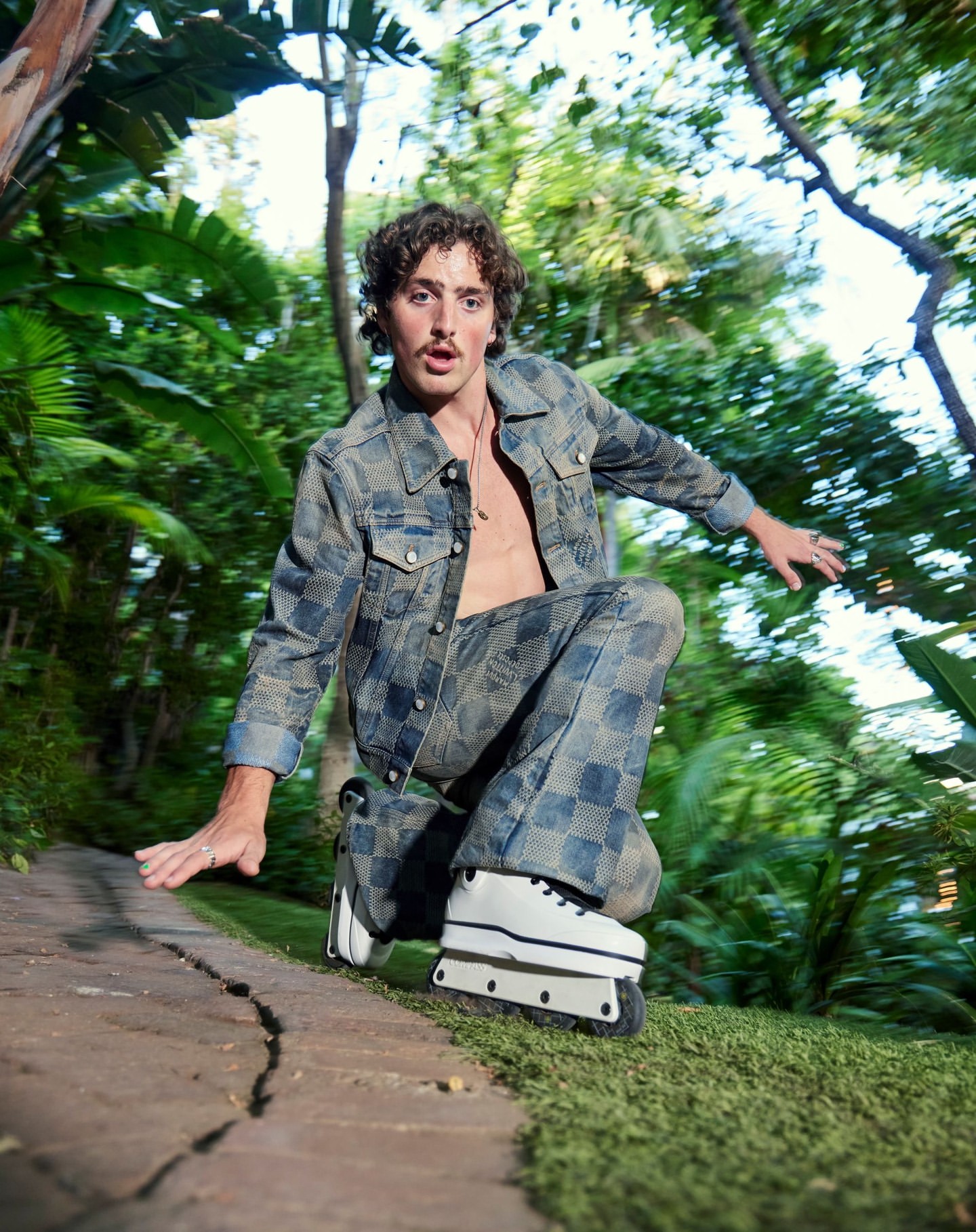 A young adult male on roller skates, wearing a patterned denim jacket and pants, kneels on a stone path surrounded by green foliage.