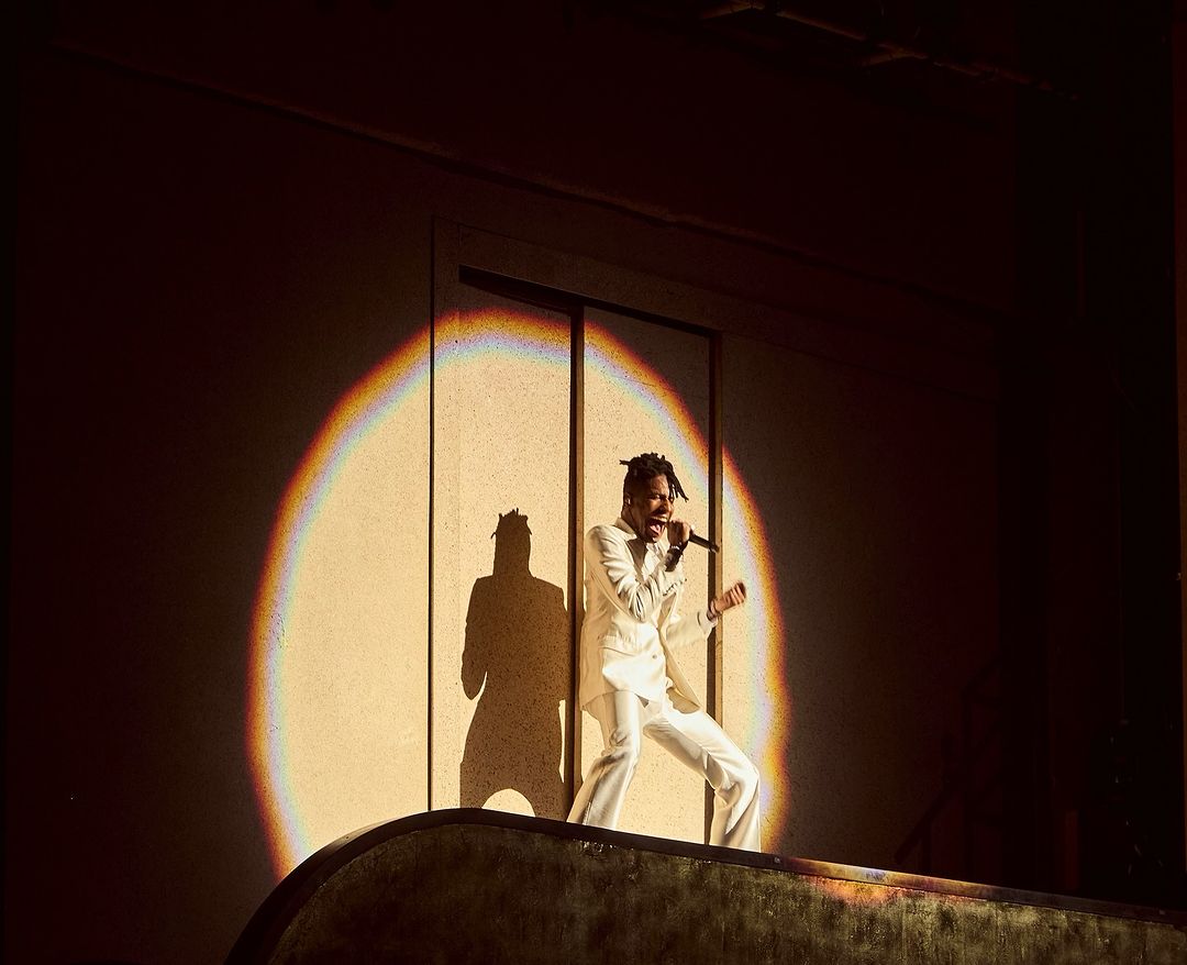A singer in a white suit performs on a stage, illuminated by a spotlight against a dark background, creating a dramatic visual.