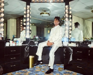 A man in a cream suit sitting on a dressing room counter between illuminated vanity mirrors, photographed by Jen Rosenstein.