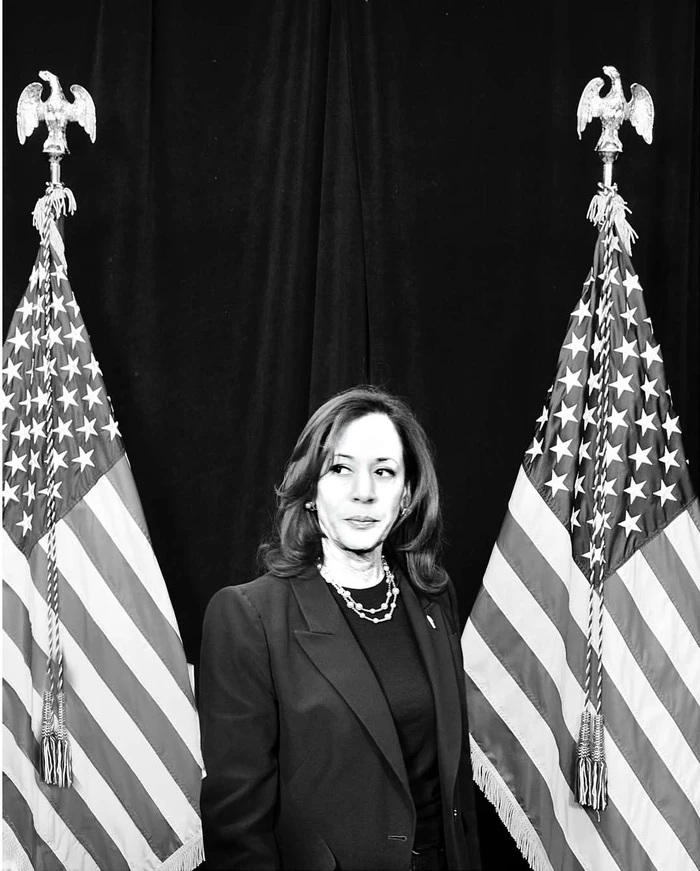 Kamala Harris wearing a dark blazer and pearl necklace standing between two American flags in monochrome