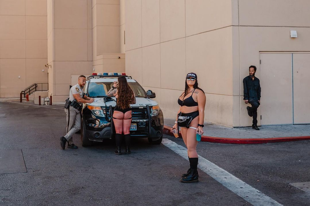 Three women in costume pose next to a police car on a city street, while a man leans against a building in the background.