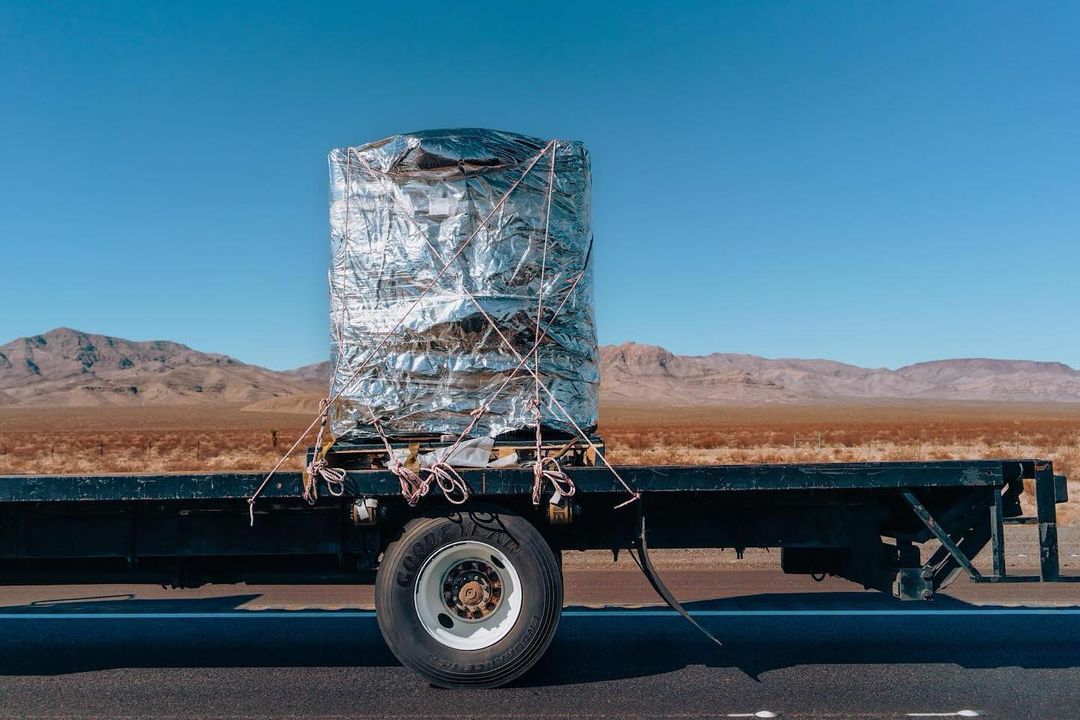 Large cylindrical object, wrapped in reflective material and secured with ropes, loaded on a black flatbed trailer against a desert background.