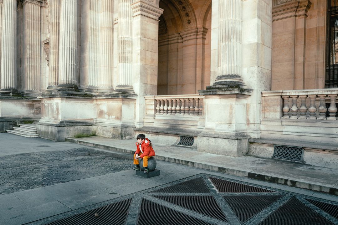 Boy in orange jacket sits on stone sphere in front of monumental architecture with columns and arches in an urban environment.