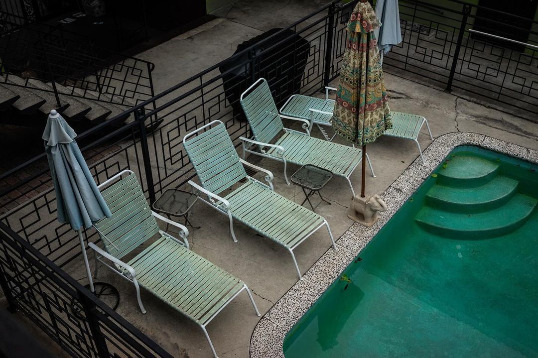 Aerial view of four pool loungers beside a bright turquoise pool, plus two colorful umbrellas on a concrete patio with iron railings.