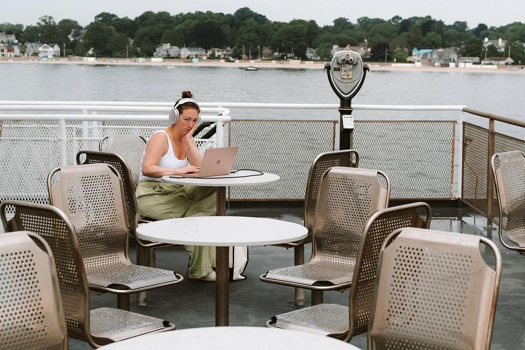 Woman with headphones works on a laptop at an outdoor table on a boat near the shoreline with houses and trees on an overcast day.