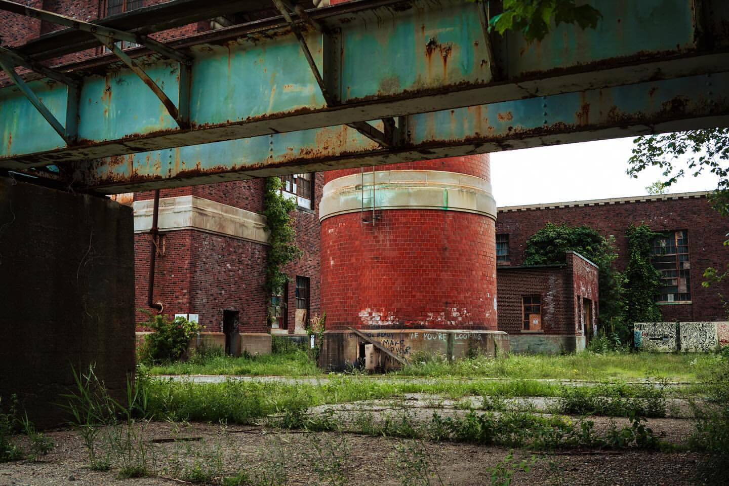 Exterior of a brick industrial building with a cylindrical tower and rusty metal structure in the foreground, overgrown with grass.