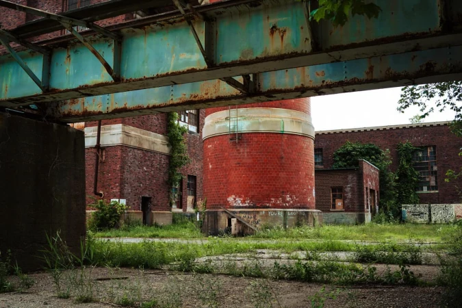 Exterior of a brick industrial building with a cylindrical tower and rusty metal structure in the foreground, overgrown with grass.