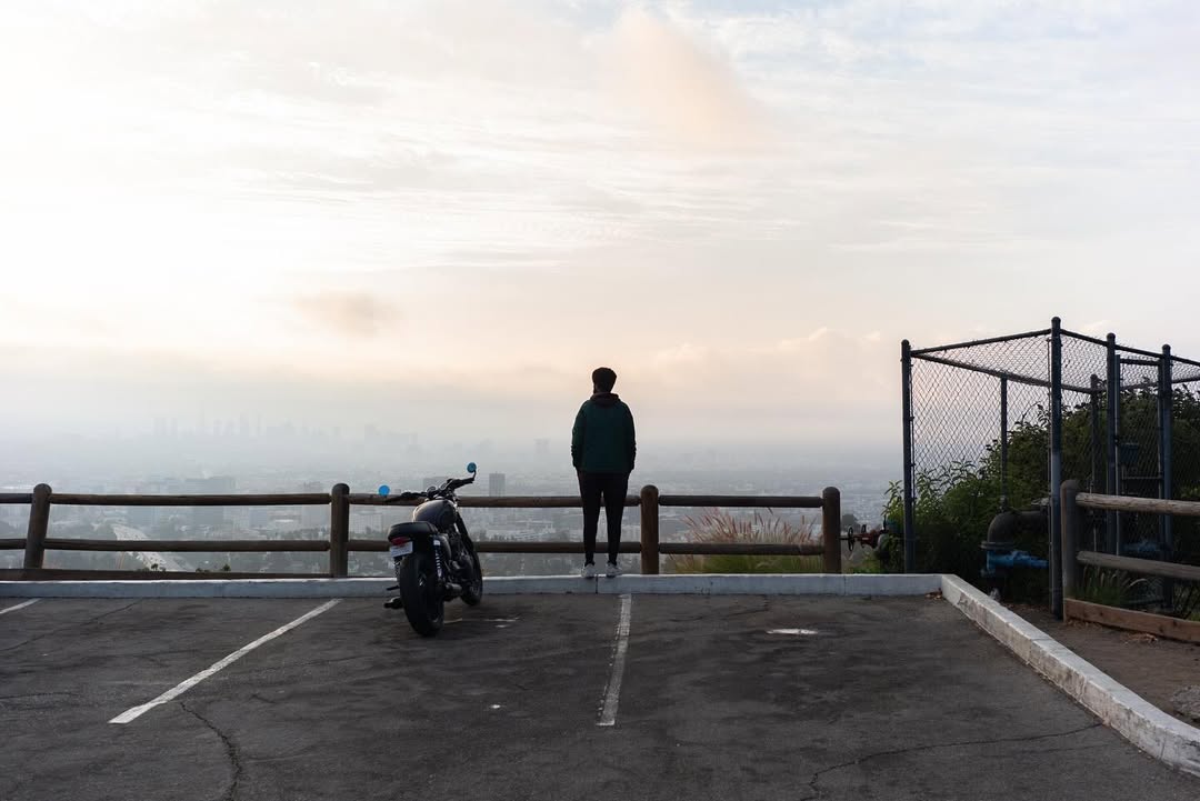 A person stands near a motorcycle on a parking lot overlook with a cityscape shrouded in fog and a bright, overcast sky at dawn.