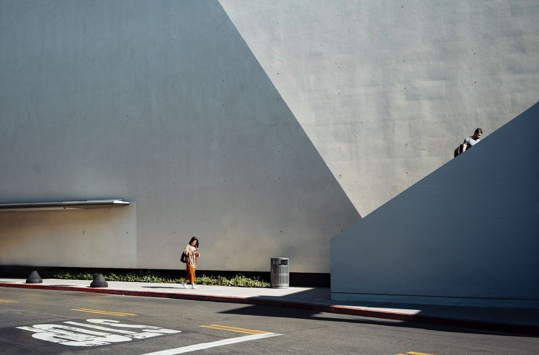 Two pedestrians on a city sidewalk alongside a minimalist, geometric building with a concrete wall and angular shadow patterns.