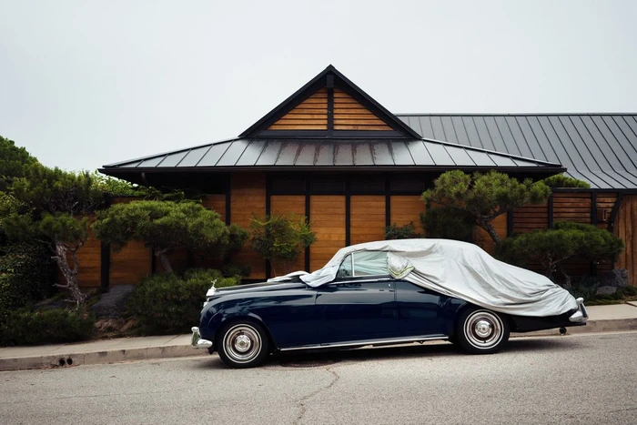 Classic dark blue car covered with a light grey sheet parked in front of a modern wood-paneled building with mature pine trees.