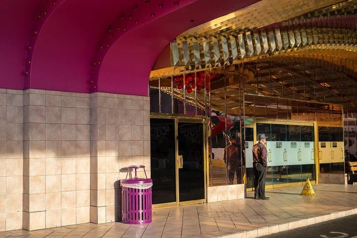 A man wearing a hat stands outside a building entrance with vibrant pink arches and gold mirrored paneling.