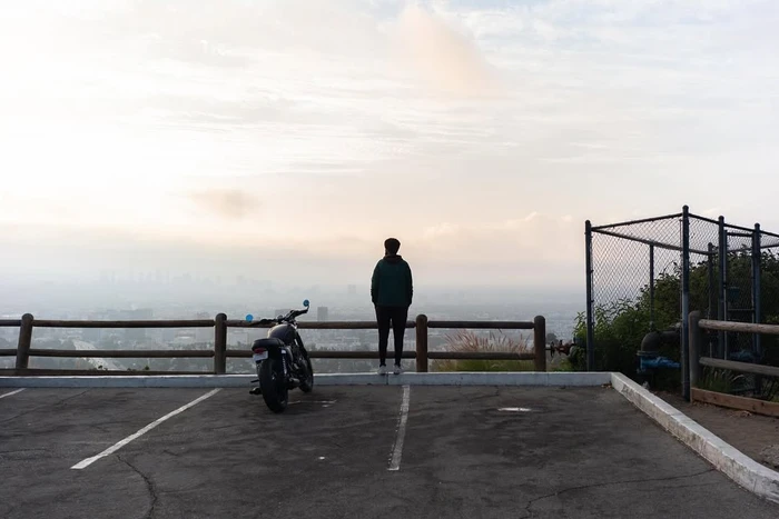 A person standing at a wooden fence overlooking a hazy cityscape, with a motorcycle parked in an asphalt parking lot nearby.