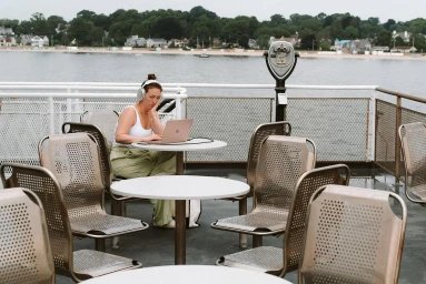 Woman wearing headphones seated at a table on a ferry deck using a laptop, photographed by Renny Maslow.
