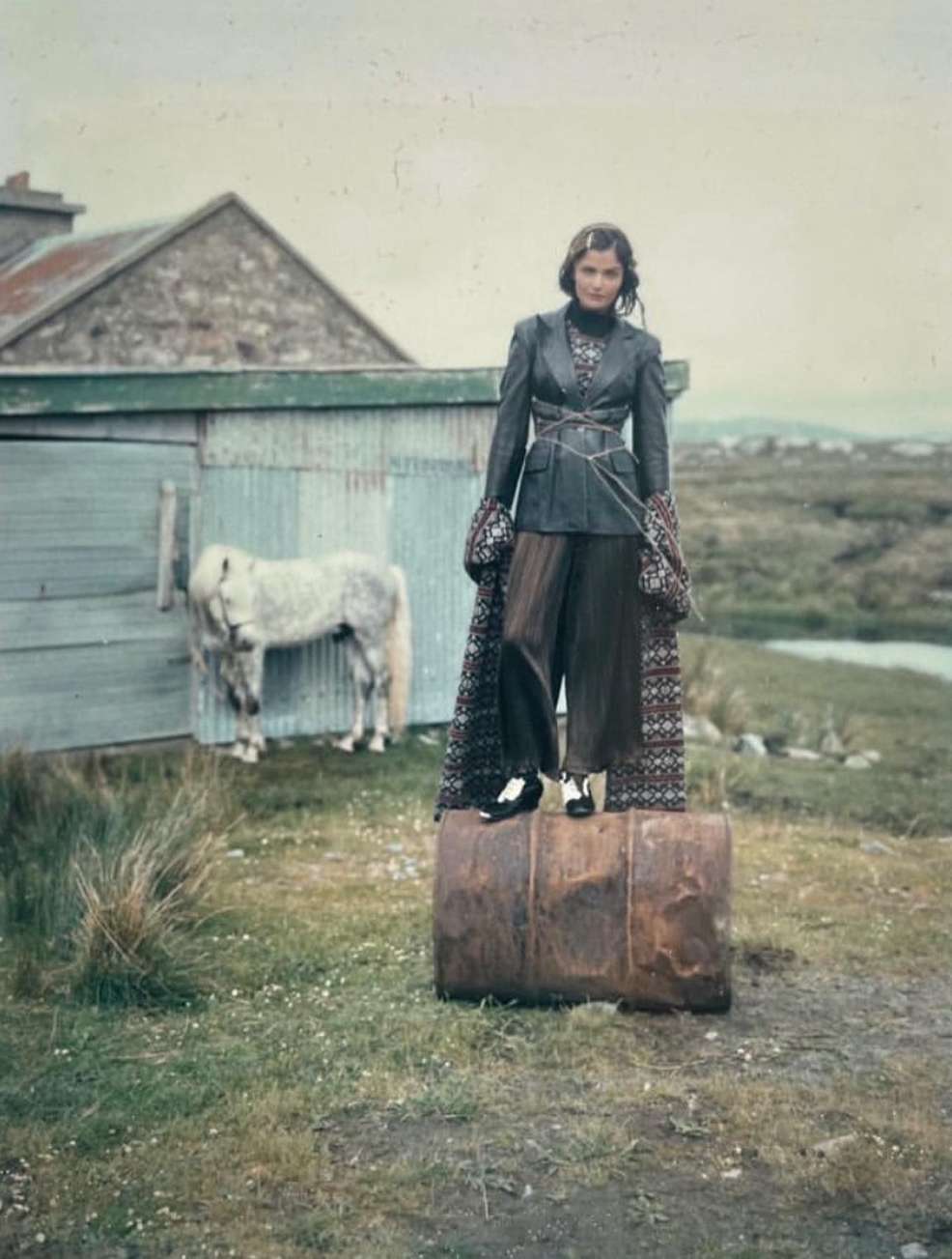 Fashionable woman in jacket and wide-leg pants standing on barrel outdoors with a white pony, stone shed, and landscape background.