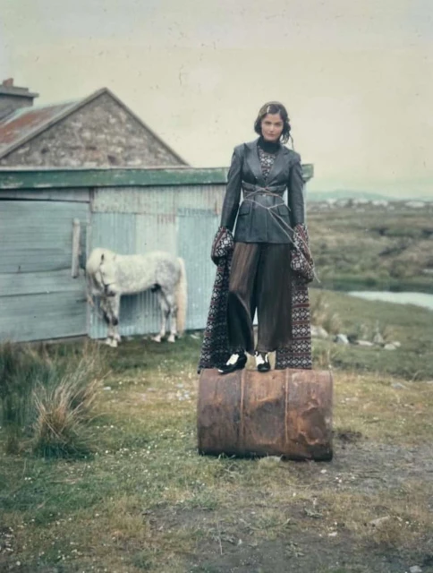 Fashionable woman in jacket and wide-leg pants standing on barrel outdoors with a white pony, stone shed, and landscape background.