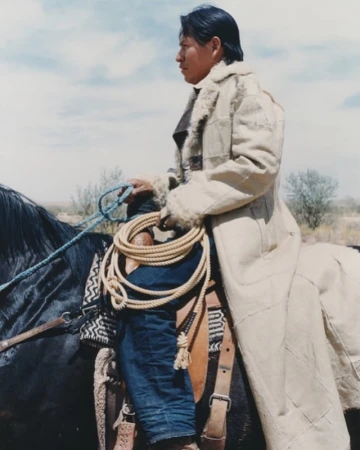 An Indigenous man rides a black horse in a light beige coat, blue jeans and a rope across his lap against a dry brush landscape.