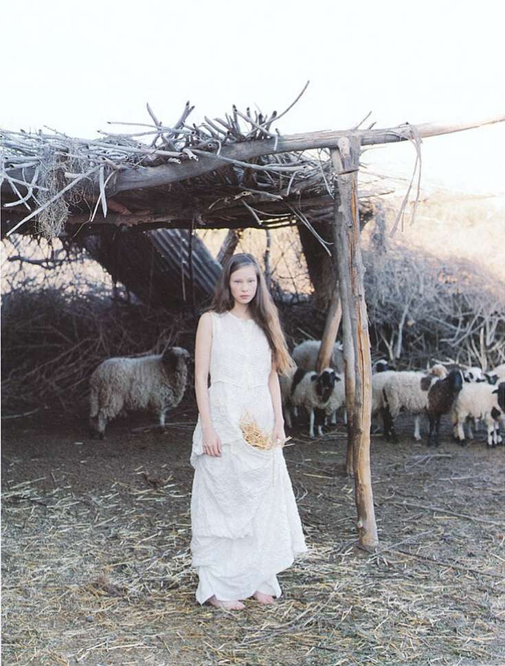 Young woman in white dress stands in a farmyard with a flock of sheep and a wooden shelter, creating a rustic, pastoral aesthetic.
