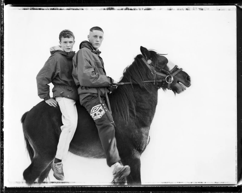 Two young men ride a brown horse against a bright background, wearing jackets and sneakers in a monochromatic, timeless composition.