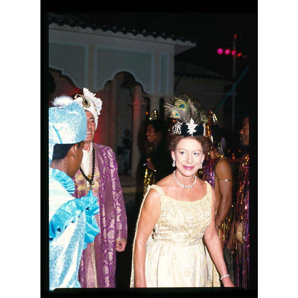 Elegant woman in a gold gown and jeweled headdress attends an event with other formally dressed guests near a building at night.
