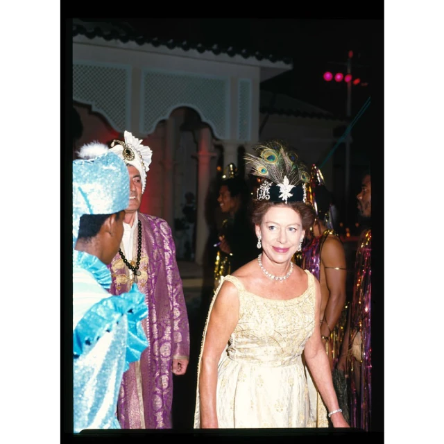 Elegant woman in a gold gown and jeweled headdress attends an event with other formally dressed guests near a building at night.