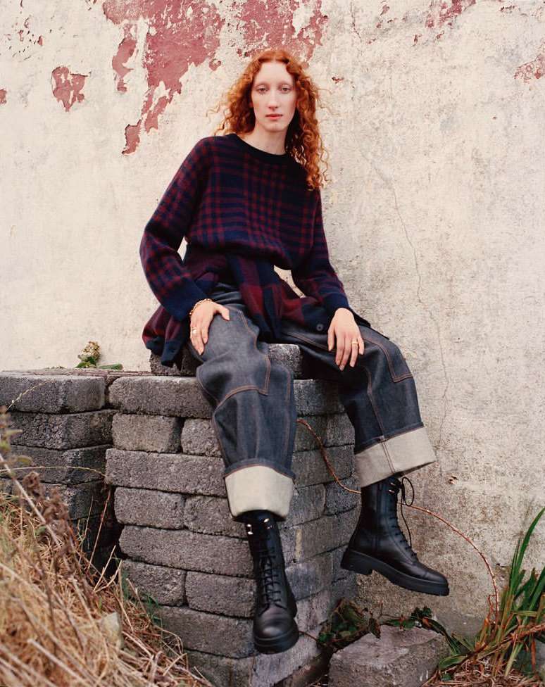 A red-haired woman in a checkered sweater and cuffed jeans sits on a stone structure in front of a textured wall, wearing black boots.