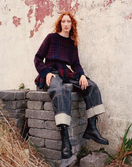 A red-haired woman in a checkered sweater and cuffed jeans sits on a stone structure in front of a textured wall, wearing black boots.
