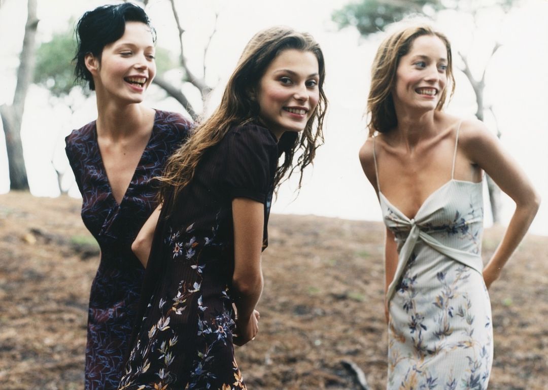 Three women in floral dresses stand closely together in a grassy outdoor setting with trees in the background, all smiling and relaxed.